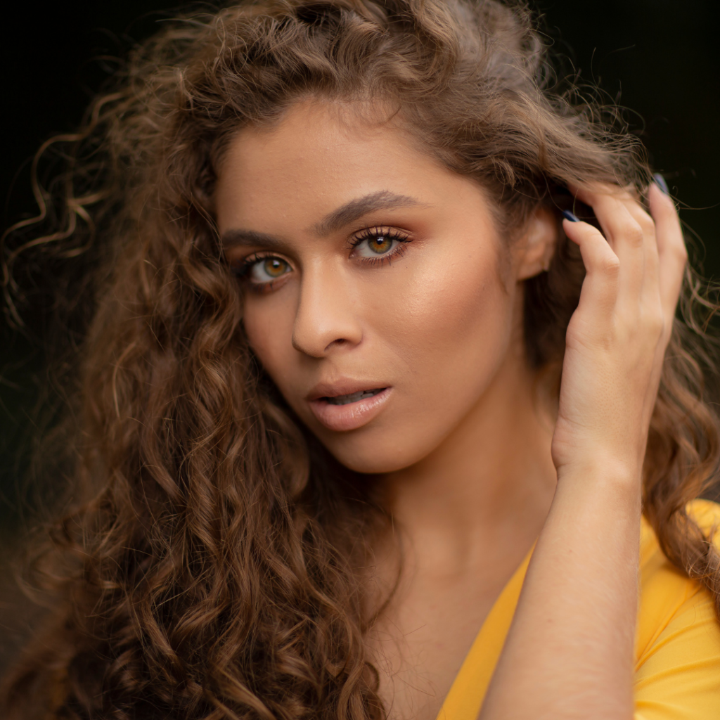 A woman with curly hair wearing yellow and lifting her hair below her left ear